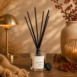 May include: A clear glass reed diffuser bottle with a white label reading Adelica Candle Co and golden glow 4 oz reed diffuser. Six thin black reeds extend from the top of the bottle, and a small black circular cap sits beside the glass base.
