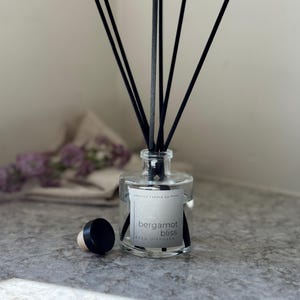 May include: A clear glass reed diffuser with black reeds and a black cap. The label reads "bergamot bliss REED DIFFUSER". The diffuser is on a gray surface. The product is from "Arelia Candle Co."