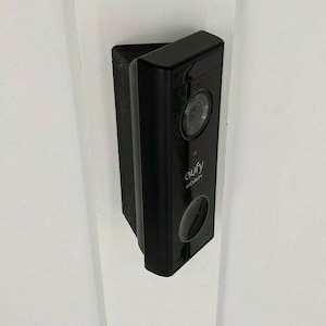 May include: A black Eufy Security video doorbell mounted on a white door frame. The doorbell has a camera lens, a motion sensor, and the Eufy logo. The doorbell is rectangular and has rounded edges.