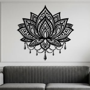 Puede incluir: Arte mural de flor de loto de metal negro. El intrincado diseño presenta pétalos detallados y acentos colgantes. La obra de arte está montada en una pared blanca sobre un sofá gris, con una planta en maceta en la esquina.