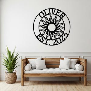Può includere: Opera d'arte da parete in metallo nero con un design a sole, il nome "OLIVER" e "EST. 2023" in una cornice circolare. Il sole ha raggi stilizzati. Montato su un muro bianco sopra un divano in legno.