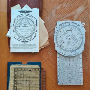 Puede incluir: Un surtido de herramientas de navegación aérea vintage, que incluyen una regla de cálculo circular y una computadora de vuelo, con el texto "NCI FLIGHT COMPUTER". Las herramientas están sobre una superficie de madera, con un estuche de cuero marrón y varios papeles.