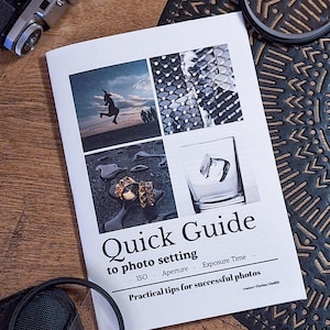 Op de afbeelding: Een wit boekje met de titel "Snelle gids voor foto-instellingen" en de tekst "ISO, Diafragma, Belichtingstijd" en "Praktische tips voor succesvolle foto's". Het boekje is open op een pagina met vier afbeeldingen: een persoon die in de lucht springt, een zwart-witafbeelding van een gezicht, een close-up van een gouden ring en een glas met ijsblokjes.