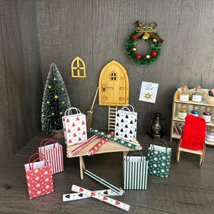 Könnte beinhalten: Miniatur-Weihnachtsdekorationen, darunter Geschenktüten mit Stern- und Baummustern, Geschenkpapierrollen, ein kleiner Weihnachtsbaum und ein Kranz. Eine Holztür, ein Fenster und ein Regal vervollständigen die festliche Szene.