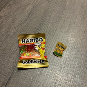 Könnte beinhalten: Zwei Packungen Haribo Goldbären Gummibärchen. Eine volle Packung und eine kleine, zerrissene Packung. Die volle Packung ist goldfarben mit dem Haribo-Logo und einem Cartoon-Bären. Die kleine Packung ist ebenfalls goldfarben mit dem Haribo-Logo.