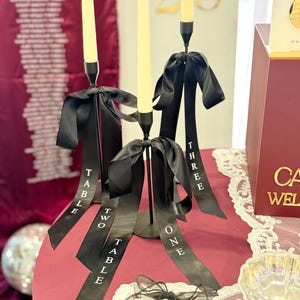 May include: Three black metal candle holders with cream candles and black ribbon accents. Each ribbon is labelled with "TABLE ONE", "TABLE TWO", and "TABLE THREE" in white text. The table is covered with a red cloth.
