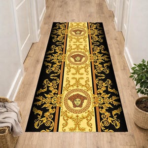 Puede incluir: Una alfombra rectangular con un diseño ornamental negro y dorado. La alfombra presenta un panel central amarillo con medallones de cabeza de Medusa y volutas doradas que se repiten. Los lados son negros con volutas doradas. La alfombra está colocada en un pasillo.