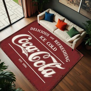 May include: A rectangular area rug featuring a distressed red background with the white Coca-Cola logo, the text "DELICIOUS and REFRESHING", "ICE COLD", and "EST. 1886". The rug is positioned on a dark wooden floor in front of a beige sofa.