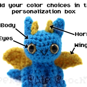 May include: A crocheted blue dragon with yellow wings and horns. The dragon has black eyes and a white mouth.