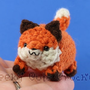 Puede incluir: Un pequeño zorro de crochet con pelaje naranja, detalles blancos y orejas marrones. El zorro está sentado en una mano con un anillo.