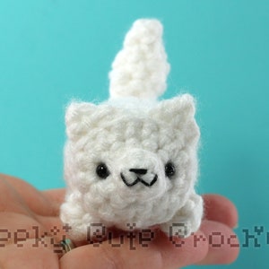 Puede incluir: Un gato blanco de ganchillo hecho a mano con ojos negros y una sonrisa cosida. El gato tiene una cola esponjosa y se sostiene en una mano. El texto "Geeky Cute Crochet" es visible en la esquina inferior izquierda.