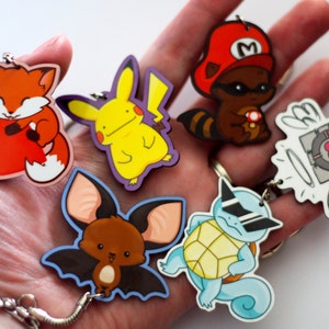 Puede incluir: Un conjunto de seis llaveros coloridos con personajes de dibujos animados. Los llaveros incluyen un zorro rojo, un Pikachu amarillo, un murci&eacute;lago marr&oacute;n, una tortuga azul con gafas de sol, un mapache con un sombrero rojo y un cubo blanco y gris con alas.