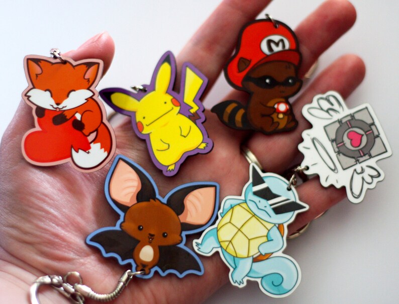 Puede incluir: Un conjunto de seis llaveros coloridos con personajes de dibujos animados. Los llaveros incluyen un zorro rojo, un Pikachu amarillo, un murci&eacute;lago marr&oacute;n, una tortuga azul con gafas de sol, un mapache con un sombrero rojo y un cubo blanco y gris con alas.