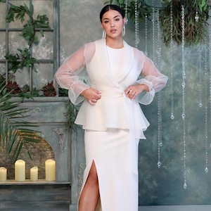 May include: A white formal dress with a matching sheer overlay jacket. The dress features a high neckline, a peplum waist, and a thigh-high slit. The jacket has sheer sleeves and a draped front. The outfit is elegant and suitable for special occasions.