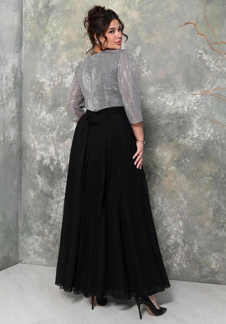 May include: A formal dress featuring a silver sequined top with three-quarter sleeves and a long, flowing black skirt. The dress has a tie at the waist and is paired with black high-heeled shoes. The background is a textured gray wall.