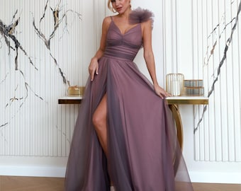 May include: A mauve-colored formal gown with a high slit and spaghetti straps. The dress features a ruffled shoulder detail and a flowing, layered skirt. The bodice is ruched, and the dress appears to be made of a soft, sheer fabric.