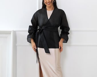 May include: A black wrap-style blouse with long, voluminous sleeves featuring gathered cuffs and a matching fabric tie at the waist, paired with a floor-length, champagne-coloured satin skirt that has a high side slit along the leg.