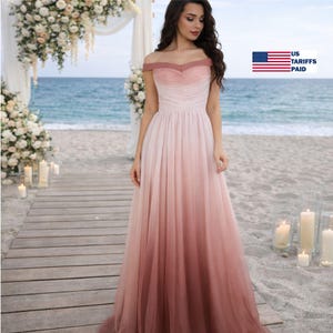May include: A floor-length, off-the-shoulder gown in a gradient of pink and mauve. The dress features a ruched bodice and a flowing skirt. The background includes a beach setting with floral decorations and the text "US TARIFFS PAID".