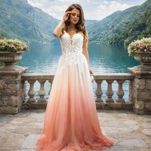 May include: A white and peach ombre wedding dress with a V-neckline and floral lace detailing. The dress has a flowing skirt and is worn outdoors with a scenic lake and mountain backdrop.