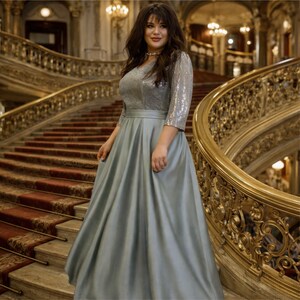 May include: A formal gown with a silver sequined bodice and a flowing, pale blue skirt. The dress has three-quarter sleeves and a v-neckline. The background features a grand staircase with ornate gold railings and red carpet.