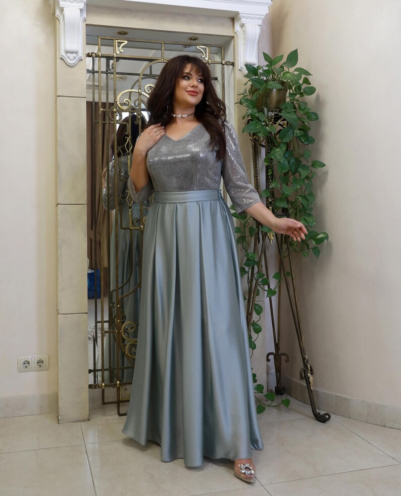 May include: A formal, floor-length gown in a soft, silvery-blue hue. The bodice features a V-neck and is embellished with sequins, while the skirt is a flowing, pleated design. The dress has three-quarter sleeves and is suitable for special occasions.
