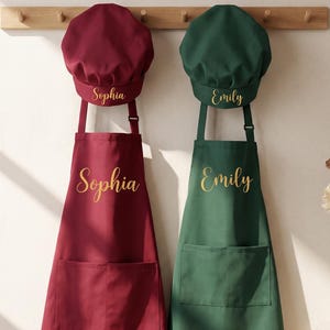 May include: A burgundy apron and chef hat set personalized with the name Sophia in gold script, alongside a dark green apron and chef hat set personalized with the name Emily in gold script, both hanging on a wooden wall rack.