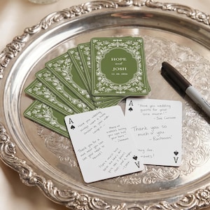 May include: A silver decorative tray holding a stack of green playing cards with white floral borders and the text Hope and Josh 05.08.2026, alongside two white playing cards with handwritten black ink messages and a black marker pen.