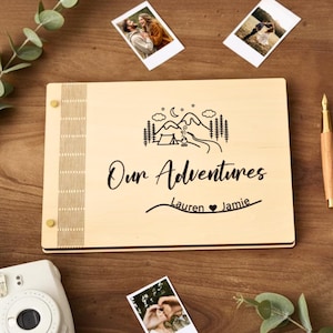 Könnte beinhalten: Ein helles Holz-Fotoalbum mit den Worten "Our Adventures" und den Namen "Lauren & Jamie" ist zu sehen. Das Cover zeigt ein Berg- und Baumdesign. Eine weiße Sofortbildkamera, Fotos und ein Stift sind ebenfalls sichtbar.