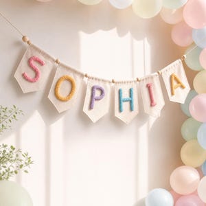 May include: A decorative banner featuring seven off-white fabric pennants suspended by a string with wooden beads. Each pennant displays a single letter in a raised, knitted texture, spelling out the name SOPHIA in pink, yellow, purple, blue, and pink.