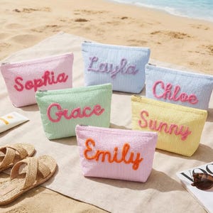 May include: A collection of personalized cosmetic bags in various pastel colors, including pink, blue, green, and yellow, each embroidered with a different name. The bags are arranged on a beach towel with sandals, sunscreen, and a magazine.