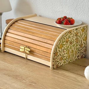 Op de afbeelding: Een houten broodtrommel met een roldeksel, horizontale latten en een goudkleurige handgreep in de vorm van een libel. De zijpanelen zijn versierd met een botanisch patroon van groene bladeren en gele vruchten op een crèmekleurige achtergrond.