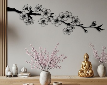 May include: A black metal wall art piece depicting a cherry blossom branch with delicate flowers. The artwork is mounted on a light gray wall, complementing the neutral tones of the room. The room features a round wooden coffee table, a gold Buddha statue, and a paper lantern.