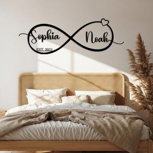 Op de afbeelding: Zwarte metalen wandkunst met een oneindigheidssymbool, de namen "Sophia" en "Noah" en het jaar "EST. 2021". Een hart is inbegrepen. Het kunstwerk wordt weergegeven boven een bed met een geweven hoofdbord en neutraal beddengoed.
