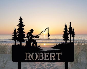 Puede incluir: Letrero de metal negro con la silueta de un pescador con caña, un pez capturado y pinos, con el nombre "ROBERT" debajo. El fondo muestra una escena de playa al atardecer.