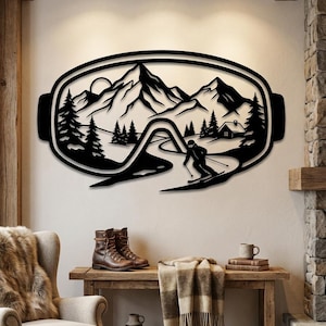 Op de afbeelding: Een zwart metalen wanddecoratie in de vorm van een skibril, met een uitgesneden ontwerp van een bergketen, dennenbomen, een kleine hut en een skiër op een helling. Het gedetailleerde silhouet toont een winterlandschap.