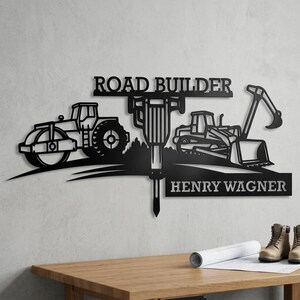 May include: Black metal road builder sign with a steamroller, jackhammer, bulldozer, and excavator. The sign reads "ROAD BUILDER" and "HENRY WAGNER". The sign is mounted on a wall.