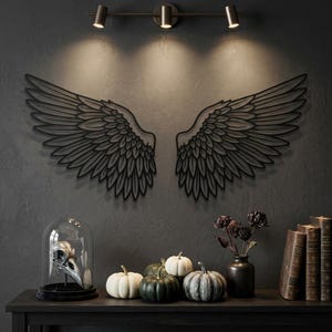 Op de afbeelding: Een zwart metalen wanddecoratie met twee symmetrische engelvleugels met gedetailleerde veeruitsnijdingen. De vleugels zijn bevestigd aan een donkergrijze muur boven een tafel met een vogelschedel onder een glazen stolp en pompoenen.