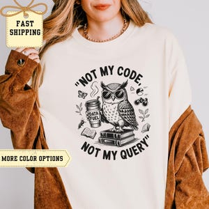 Könnte beinhalten: Cremefarbenes T-Shirt mit einem Aufdruck einer Eule mit Sonnenbrille, die eine Kaffeetasse hält und auf Büchern sitzt. Der Text lautet "NOT MY CODE, NOT MY QUERY". Das Design umfasst einen Schmetterling, ein Fernglas und Blätter.