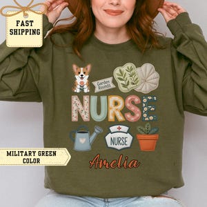 May include: Military green sweatshirt with a colorful "NURSE" design. The design includes a corgi, watering can, and potted plant. The name "Amelia" is written in cursive. The sweatshirt has the text "Garden Rounds" and "NURSE" in the design.