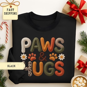 May include: Black sweatshirt with the words "PAWS & HUGS" in a colorful, stitched design. The design includes paw prints, daisies, and a leopard print accent. The sweatshirt is surrounded by holiday decorations.