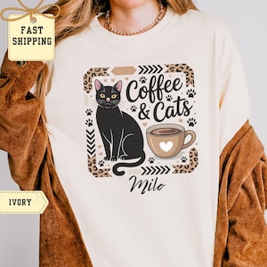 Puede incluir: Camiseta color marfil con una ilustración de gato negro con ojos amarillos, una taza de café con un corazón y el texto "Coffee & Cats" y "Milo". El diseño está enmarcado por un borde de estampado de leopardo y huellas de patas.