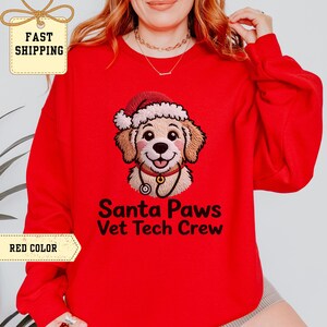 Puede incluir: Una sudadera roja con un perro de dibujos animados con un gorro de Papá Noel y un estetoscopio. El texto "Santa Paws Vet Tech Crew" está impreso debajo. Un artículo festivo para las fiestas.