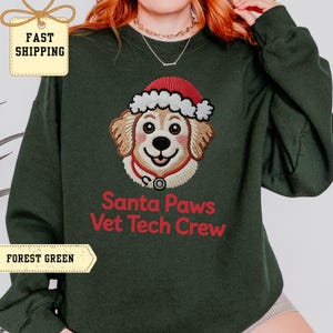 Puede incluir: Sudadera verde oscuro con un perro de dibujos animados bordado con un gorro de Papá Noel y un estetoscopio. El texto "Santa Paws Vet Tech Crew" es rojo. Un diseño festivo navideño.