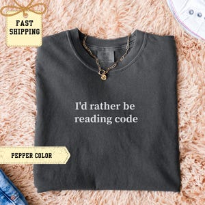 Peut inclure: T-shirt gris foncé avec l'inscription blanche "I'd rather be reading code". Le t-shirt a un col rond et des manches courtes. Un collier doré est posé sur le t-shirt. Le t-shirt est présenté sur une surface rose et moelleuse.