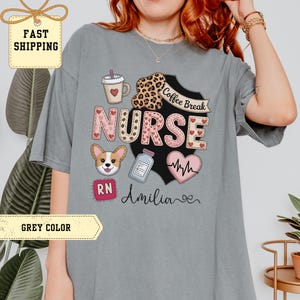 May include: A gray t-shirt featuring a colorful graphic design for nurses. The design includes the word "NURSE" with hearts, a coffee cup, a corgi, a medical vial, and a heartbeat. The text "Coffee Break" and "Amilia" are also included.