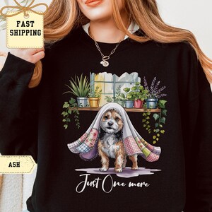 Può includere: Felpa nera con un cane sotto una coperta, sullo sfondo di una mensola con piante in vaso. La scritta "Just One more" è scritta in corsivo nella parte inferiore dell'immagine.