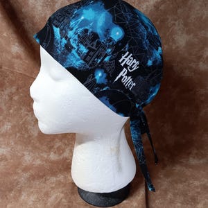 May include: A blue and black patterned head wrap featuring the text "Harry Potter" in white. The head wrap is tied at the back and displayed on a white mannequin head. The fabric has a celestial design with buildings and stars.