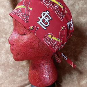 May include: Red and white patterned scrub cap featuring the St. Louis Cardinals logo and team name. The cap is tied at the back and is displayed on a red mannequin head.