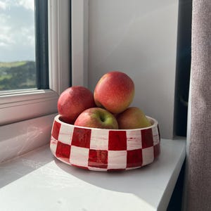 May include: A round ceramic bowl featuring a hand-painted red and white checkerboard pattern, filled with four fresh red apples. The bowl has a low profile with straight sides and a glossy finish, displaying visible brushstrokes in the paint.