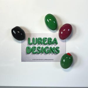 May include: A rectangular card featuring the text LUREBA DESIGNS and the URL ETSY.COM/SHOP/LUREBADESIGNS, surrounded by four smooth, oval-shaped objects: one black, one dark green, one deep red, and one green with a small red center.